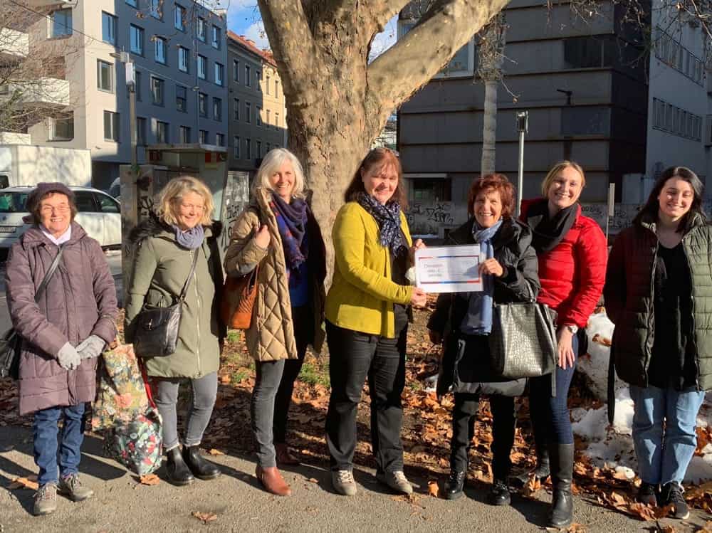 A group of women in winter coats stand outside in front of a tree.